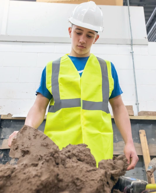 A student in a white safety helmet and high-visibility vest standing at a workstation, looking down thoughtfully at mortar and bricklaying tools during a brickwork training session. A student in a white safety helmet and high-visibility vest standing at a workstation, looking down thoughtfully at mortar and bricklaying tools during a brickwork training session.